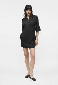 Nő áll, fekete, rövid ujjú ruhát visel cipzáros gallérral és fekete balerinacipővel, egyik kezét a zsebébe dugva pózol, fehér háttér előtt.