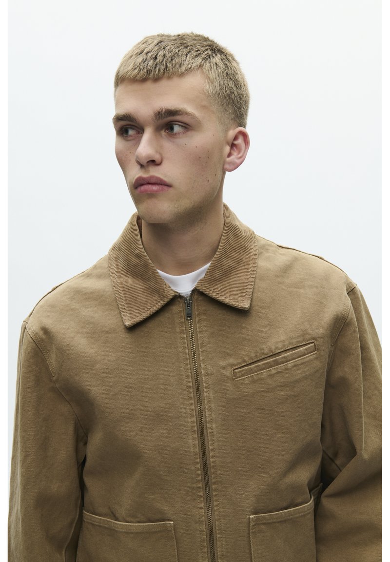 Veste en coton marron avec un col en velours côtelé, une fermeture éclair sous patte, et des poches avant. Design simple avec une finition texturée.