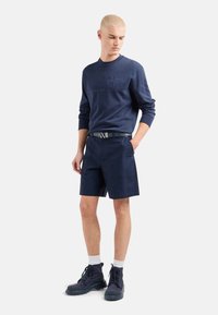 Felpa navy con logo in rilievo, abbinata a pantaloni corti navy con tasche. La calzatura è costituita da stivali scuri con suola testurizzata e calzini bianchi.