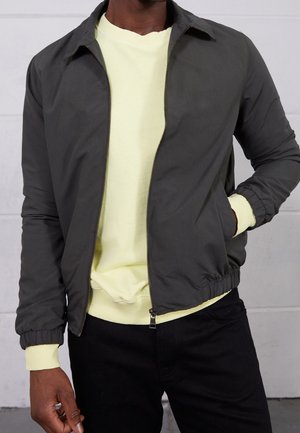 Homme portant une veste zippée gris foncé par-dessus un sweat-shirt jaune clair et un pantalon noir, une main dans la poche de la veste.