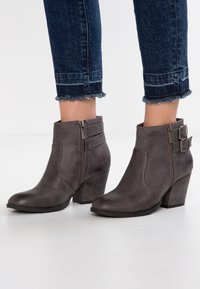 Botas de tobillo grises hechas de material sintético, con cremallera lateral, dos hebillas decorativas, puntera redondeada y diseño de tacón cuadrado.