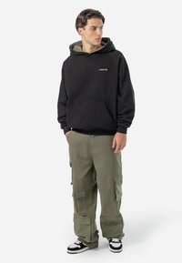 Zwarte hoodie met groene voering, voorzak en logo, gecombineerd met olijfgroene cargobroek met meerdere zakken en zwart-witte sneakers.