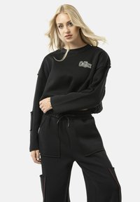 Zwart cropped sweatshirt met logo op de voorkant, met een losse pasvorm, een raw hem, en bijpassende zwarte broek met zakken en rode stiksels.