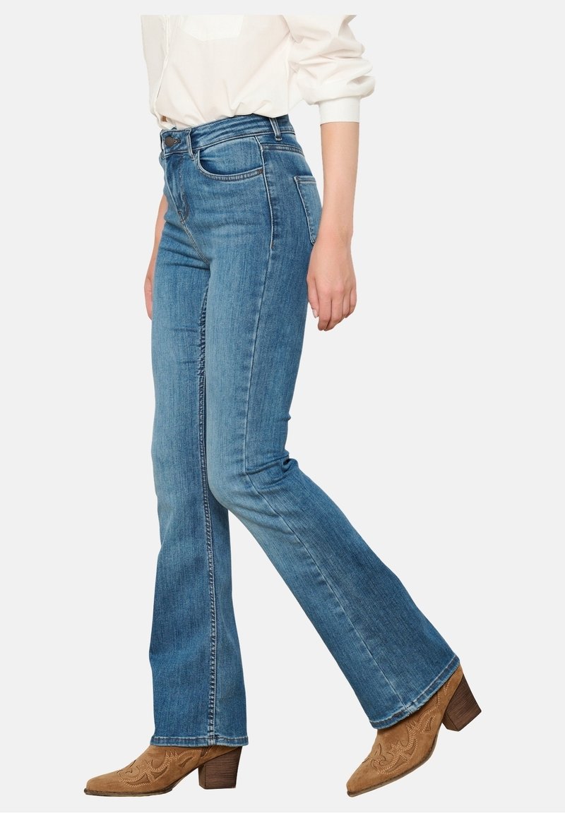 Personne portant un jean bootcut taille haute bleu, une chemise blanche à manches longues et des bottines marron brodées à talons, marchant vers la droite.