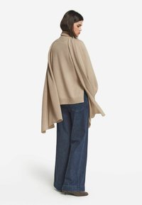Beige strikket poncho med høj krave, løst fit og draperede ærmer, parret med brede blå jeans og brune fodtøj.