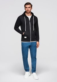 Felpa nera con zip e coulisse bianche, tasca frontale e etichetta con logo, abbinata a jeans in denim blu e scarpe da ginnastica bianche.