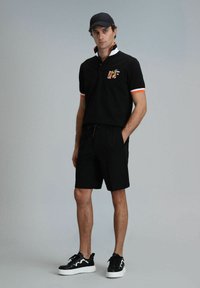 Zwart poloshirt met oranje en witte kraagaccenten; logo op de borst. Gecombineerd met zwarte shorts en zwarte sneakers met witte zolen.