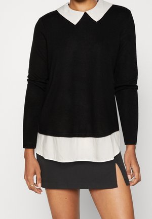 Musta pitkähihainen paita, jossa on valkoinen kaulus, kerrostettuna valkoisen paidan helman päälle, yhdistettynä mustaan minihameeseen, jossa on sivuvaō.