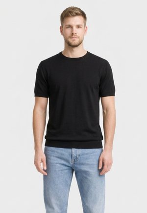 Giovane uomo con capelli corti che indossa una maglietta nera a maniche corte semplice e jeans azzurri, in piedi davanti a uno sfondo neutro.