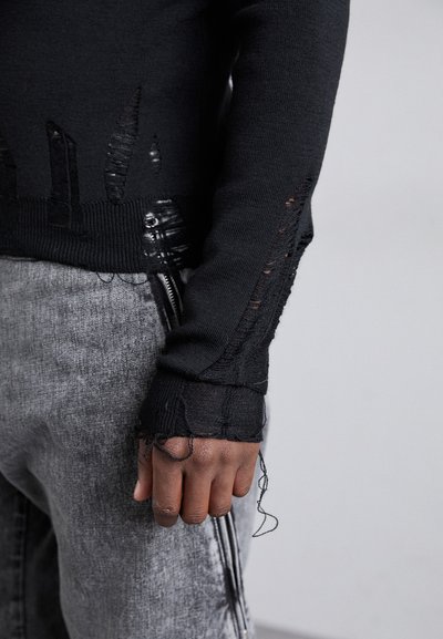 Μαύρο πλεκτό πουλόβερ με φθαρμένο σχέδιο, με τριμμένες άκρες. Γκρίζα τζιν με λεπτομέρεια πλευρικού φερμουάρ. Γενικά, έχει λεία υφή.