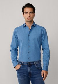 Lichtblauwe denimshirt met lange mouwen, een button-down kraag en bruine knopen. Subtiele logodetail op de borst. Gecombineerd met donkere jeans.