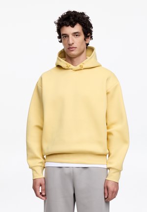 Młody mężczyzna o krótkich kręconych włosach, ubrany w bladożółtą bluzę z kapturem i jasnoszare spodnie, stojący na białym tle.