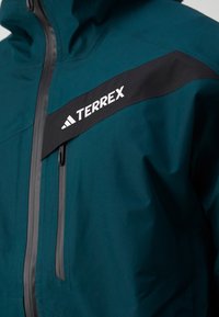 Teal outdoor jacket with a black diagonal stripe. Features a zippered chest pocket and the "TERREX" logo prominently displayed in white.