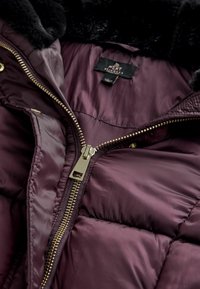 Bordeaux pufferjakke med en luksuriøs sort pelskrave. Har en guld lynlås og kviltet tekstur, der afslører en satinlignende indvendig foring.