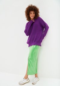 Maglione oversize in maglia viola con maniche lunghe, abbinato a una maxi gonna verde lucida con spacco laterale e sneakers nei toni neutri.