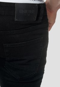 Schwarze Denim-Jeans mit einem Leder-Patch mit der Aufschrift "INDICODE" am Bund und einem ausgefallenen Design der Gesäßtasche. Glatte Textur.