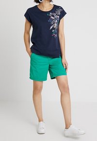 Camisa de manga corta azul marino con bordado floral en el lado izquierdo, combinada con pantalones cortos verdes. Modelo luciendo zapatillas blancas, de pie contra un fondo liso.