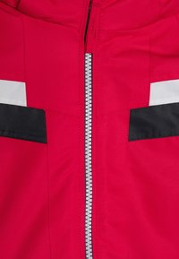 Rote Jacke mit einem zentralen silbernen Reißverschluss, versehen mit Schwarz- und Weißakzenten auf der Vorderseite. Texturiertes Gewebe mit einer glatten Oberfläche.