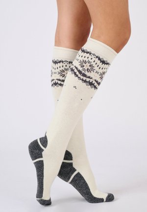 Benedenbenen met kniekousen in crèmekleur met zwarte sneeuwvlokpatronen en donkergrijze hielen en tenen, gekruist tegen een effen witte achtergrond.