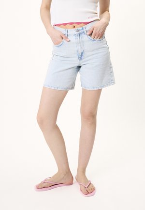 Person wearing light blue denim shorts, white cropped top, and pink flip-flops, standing with hands in pockets on plain background.