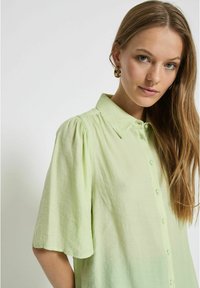 Camicia verde chiaro a maniche corte con apertura a bottoni, tessuto strutturato e sottili pieghe sulle spalle.