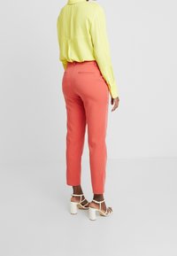 Pantalon corail à la coupe ajustée et à la longueur cheville, associé à une chemise jaune vif et des sandales blanches à talons carrés. Texture de tissu lisse.