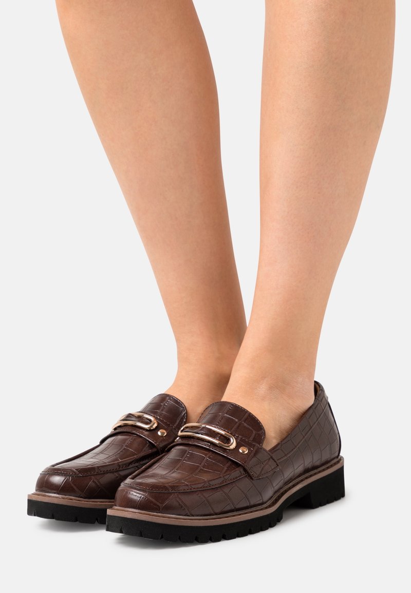 Mocassins marron à motif crocodile avec détail de boucle dorée portés sur des jambes nues sur un fond clair uni.
