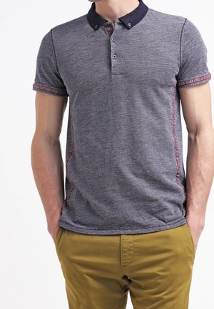 Mann trägt ein eng anliegendes, kurzärmeliges graues Polohemd mit dunklem Kragen und senfgelben Hosen, Hände in den Taschen, vor weißem Hintergrund.