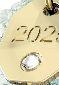 Etiqueta dorada grabada con "2023" y un acento de cristal transparente redondo, contra un fondo de cristales azules texturizados.