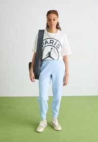Un pantalon de survêtement bleu clair associé à un t-shirt blanc avec un logo noir de Paris, complété par des baskets beige et un sac à bandoulière gris.