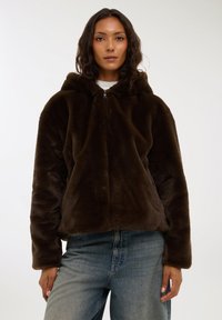 Felpa con cappuccio in pelliccia sintetica marrone scuro, con zip frontale, grande cappuccio e maniche lunghe, abbinata a jeans di denim azzurro. Texture morbida.