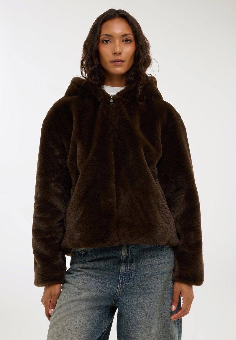 Felpa con cappuccio in pelliccia sintetica marrone scuro, con zip frontale, grande cappuccio e maniche lunghe, abbinata a jeans di denim azzurro. Texture morbida.