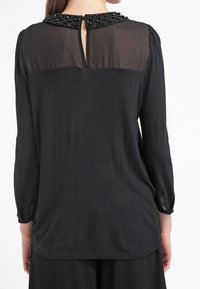 Blusa nera a maniche lunghe con una parte superiore trasparente e colletto decorato. Presenta una chiusura con bottone sul retro e polsini elasticizzati. Texture liscia.