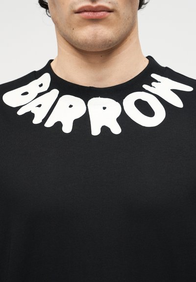 Κοντινή λήψη ενός ατόμου που φοράει μαύρο πουκάμισο με μεγάλα λευκά γράμματα που σχηματίζουν τη λέξη "BARROW" γύρω από τον λαιμό.