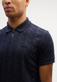 Mornarska polo majica s teksturiranim geometrijskim uzorkom, ovratnikom na zip, kratkim rukavima i suptilnim logotipom. Napravljena od glatkog materijala.