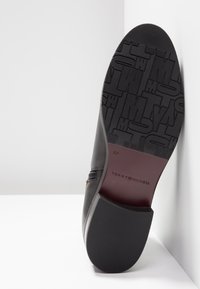 Semelle de chaussure noire avec motif texturé du logo Tommy Hilfiger, taille 37, et talon lisse, appuyée contre un mur blanc.