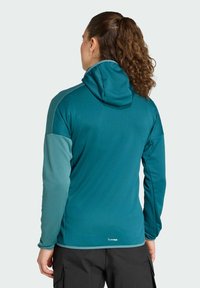 Tyrkysová kapucňová bunda s kontrastnými svetlo tyrkysovými rukávmi, dlhými rukávmi a hladkou textúrou. Na zadnom leme je umiestnené logo.