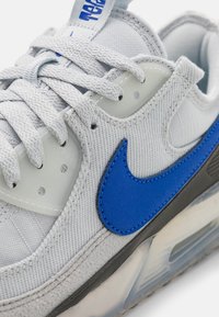 Ljusgrå sneakers med en blå läderswoosh, nätstruktur och en transparent luftkuddssula. Har grå accenter och texturerade skosnören.