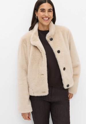 Donna che indossa una giacca beige pelosa con bottoni sopra una maglietta scura e pantaloni scuri, sorridente con capelli lunghi e scuri e orecchini a cerchio.