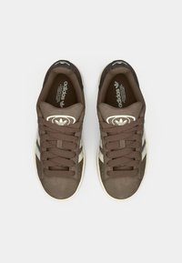 Baskets marron avec une tige en daim, bout rond, rayures blanches et fermeture à lacets. Elles présentent un talon noir texturé et une semelle intérieure de marque.