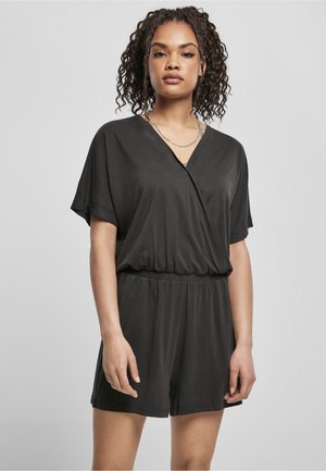 Femme aux cheveux bouclés portant une combinaison noire à manches courtes avec un col en V et une taille élastique, debout devant un fond gris clair.