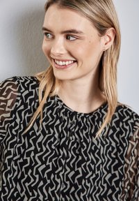 Schwarze Bluse mit abstraktem grauen Muster, durchsichtigen Ärmeln, gefälteltem Ausschnitt und glatter Textur. Goldene Creolen setzen einen metallischen Akzent.