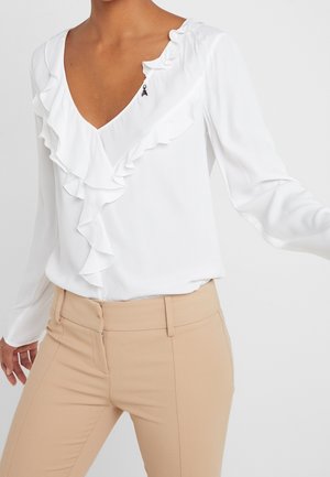 Femme portant un chemisier blanc à manches longues avec un col en V volanté et un pantalon beige ajusté sur un fond uni.