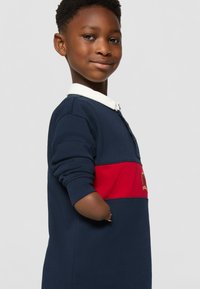 Granatowa polo z długim rękawem, z białym kołnierzykiem i czerwoną poziomą paskiem. Ma małe logo i ściągacze przy rękawach.