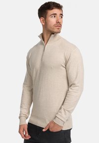 Beiger Pullover mit Reißverschluss am Hals, langen Ärmeln, gerippten Bündchen und Saum, aus weichem Stoff mit einem subtilen Strukturmuster.