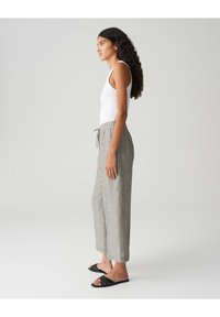 Lichtgrijze linnen broek met een ontspannen pasvorm, wijde pijpen en een elastische tailleband met een trekkoord. Gecombineerd met zwarte instapsandalen.