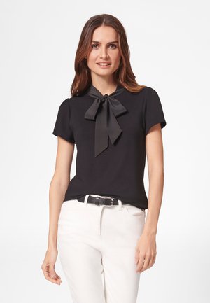 Femme portant un chemisier noir à manches courtes avec un grand col nœud papillon et un pantalon blanc avec une ceinture noire, debout sur un fond blanc.
