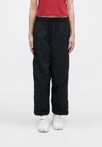 Άτομο που φοράει φαρδιά μαύρα παντελόνια, λευκά παπούτσια με χωριστό δάκτυλο και ένα κόκκινο cropped τοπ, στέκεται μπροστά σε ένα απλό λευκό φόντο.