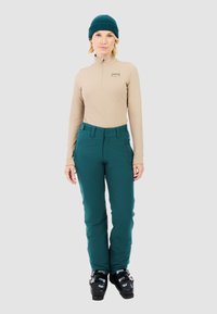 Beige zip-up basislaag top met lange mouwen, gecombineerd met teal skibroeken en zwarte skijassen, tegen een neutrale grijze achtergrond.