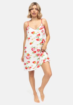 Kobieta o blond włosach, ubrana w biały halkowy sukienkę z czerwonym nadrukiem truskawek i kwiatów, stojąca boso na jednolitym tle.